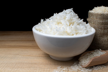 White ceramic cup with cooked rice Resting on a wooden table with a bag of rice grains and a wooden spoon, with a black backdrop.