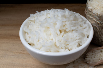 White ceramic cup with cooked rice Resting on a wooden table with a bag of rice grains and a wooden spoon, with a black backdrop.
