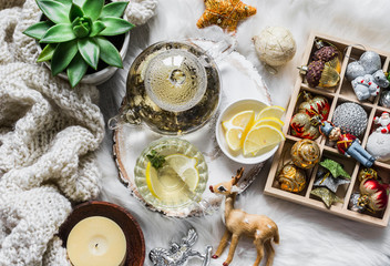 Glass teapot with green tea, flower, candle, knitted plaid and box of christmas decorations on a fluffy carpet on a light background. Christmas background