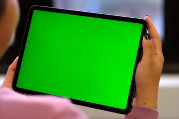 Over shoulder of woman holding green screen tablet device near window. Blur background