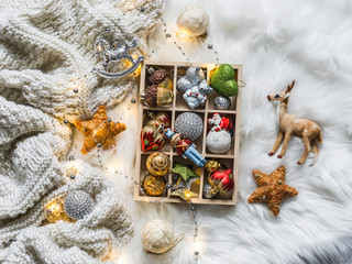 Knitted plaid, box of christmas toys, light garland on fluffy carpet, top view. Christmas background