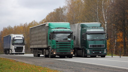 Fototapeta na wymiar Transportation logistics, European green semi trucks with awning trailers move left to right on asphalt country road on autumn day, front side wide view