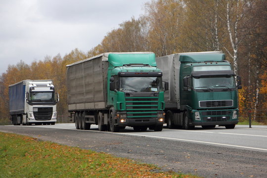 European Green Semi Trucks With Awning Trailers Overtaking On Asphalt Country Road On Autumn Day, Front Side View, Transport Logistics, Goods Delivery