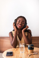 Smiling black girl listening to music on smartphone at cafe. She drinks coffee at reusable cup and singing the song.