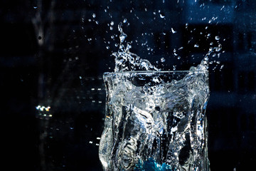 The water in the glass splashes in different directions. A glass with a crooked distorted glass with a blue ice cube falling into it, splashes of water in different directions, drops in the air