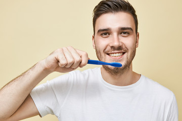 Close up image of positive young unshaven male holding plastic toothbrush while brushing teet in bathroom in front of mirror, taking care of dental hygiene, having pleased facial expression