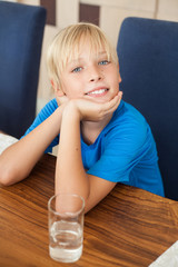 cute smiling blond teenager guy sitting in a room at a wooden table with a glass of clean drinking water