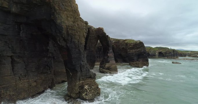Aerial: Drone approaching rock arch formations by waves splashing in sea at As Cathedrals beach against cloudy sky - Playa de las Cathedrals, Spain
