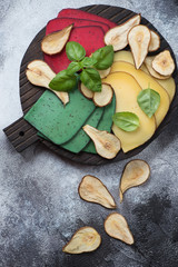 Various types of sliced cheese with pear fruit chips on a black wooden serving board. Above view over grey stone background