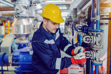 Dedicated hardworking worker in protective working clothes and with helmet on head screwing valve...