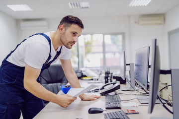 Dedicated hardworking factory worker standing in control room holding documents and looking at monitor.