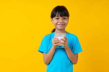 Happy Thai kid holding glass of milk isolated, young Asian girl drinking milk for strong health on yellow background