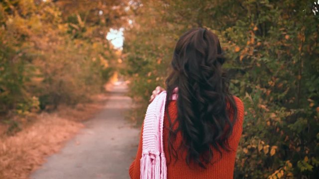 young beauty brunette woman walks along the road in orange sweater and wears a pink scarf, shot from a back, rear view. Fashionable, glamorous girl. Background autumn nature, sunny day. Yellow trees