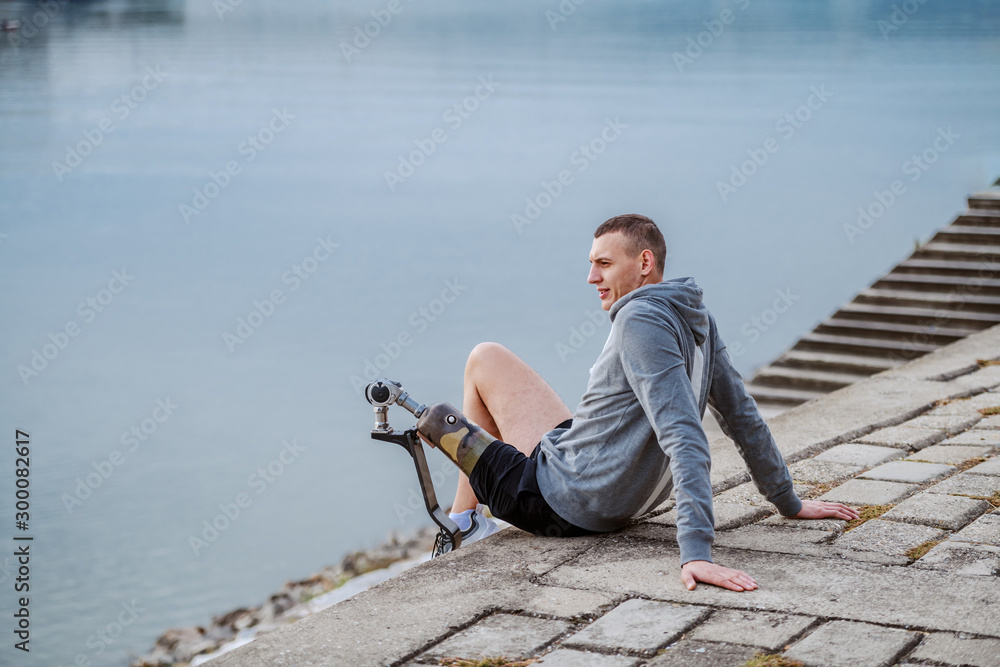 Wall mural Side view of caucasian handicapped sportsman in sportswear and with artificial leg sitting on quay and looking at river. - Wall murals
