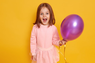 Fototapeta na wymiar Portrait of surprised little girl with dark staright hair standing over yellow studio background beautiful clothes, holding purple ballon in hands and looking at camera joyfully. Holiday concept.