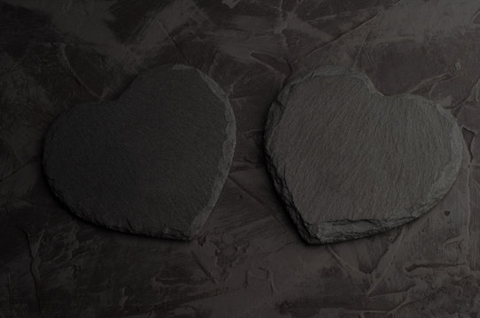 Two coasters made of black slate in the shape of a heart on a dark stucco background. Copy space