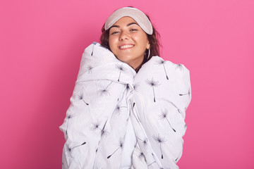 Close up portrait of woman with sleeping mask on her forehead, wrapped white blanket with dandelion, isolated over pink studio background, looking smiling at camera, feels happy, enjoying morning time