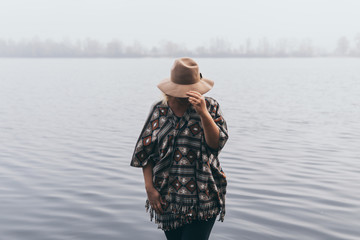 Woman in hat and poncho standing on the river bank and looking towards autumn forest covered with...