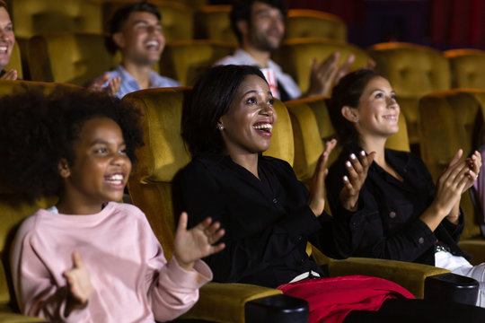 Excited black woman and girl audience smiling and clapping hands while sitting amidst people after movie in cinema