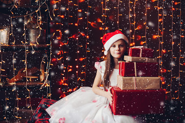 Portrait of cute girl in white dress and red Santa s hat holding many Christmas presents against...