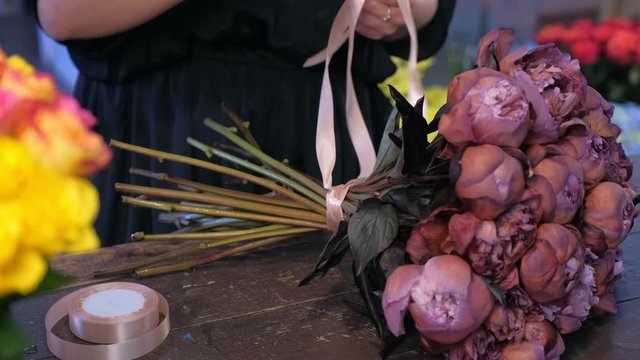 Floral business concept. Professional woman florist ties stems by ribbons on bouquet of brown maroon color peonies in flower shop for sale. She creates bunch composition in floristic studio store.