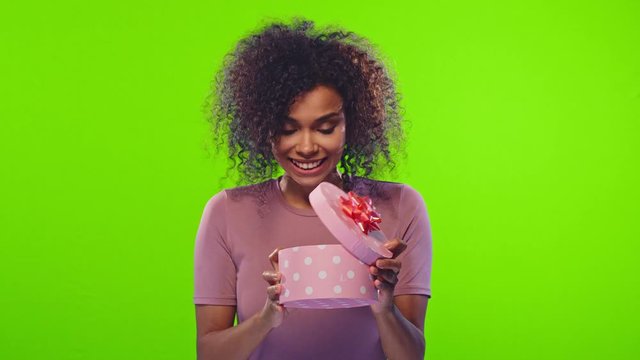 Positive emotional young pretty dark skinned woman happily opening a box with a gift and disappointed on a green background. Concept of surprise and unwarranted expectations. People and celebration