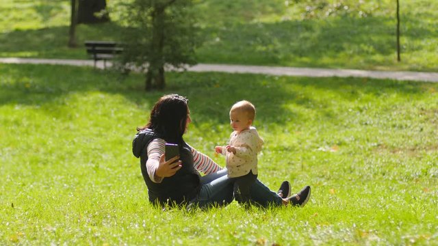 Young mother hugging toddler baby child daughter taking phone selfie picture enjoy spend time in park on a grass