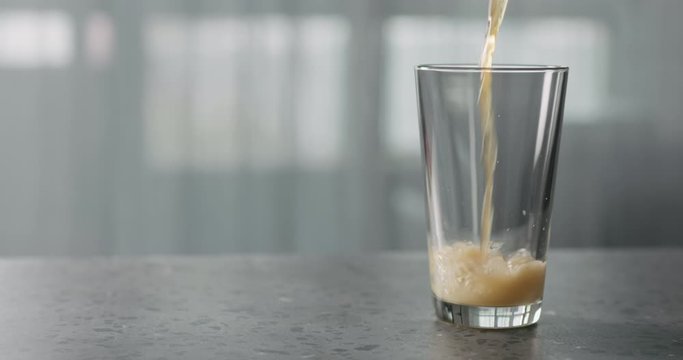 Slow motion nitro light ale beer pouring into pint glass on terrazzo countertop with window on background