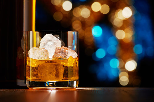 Glass of whiskey with ice and whiskey bottle on colorful Christmas lights bokeh background