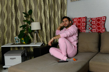 Naklejka premium Young television addict man sitting on sofa at home and watching TV using remote control, looking bored and tired, pumping someone else's comedy comedy movie or live music at night