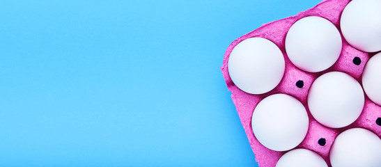 White chicken eggs in a pink carton tray box on blue background with copy space