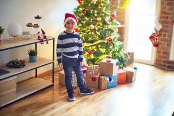 Obraz na płótnie Canvas Adorable toddler smiling happy and confident. Standing wearing santa claus hat with smile on face around christmas tree and lots of gifts at home