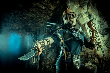 Post-apocalyptic woman in the rusty helmet with a chain and knife in hands on the dungeon...
