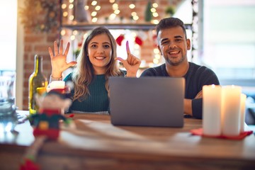 Young beautiful couple sitting using laptop around christmas decoration at home showing and...