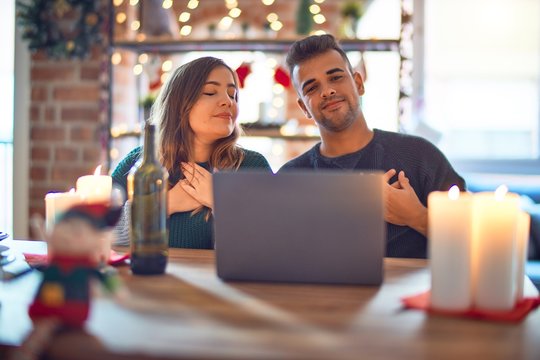Young beautiful couple sitting using laptop around christmas decoration at home smiling with hands on chest with closed eyes and grateful gesture on face. Health concept.