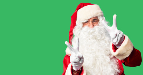 Middle age handsome man wearing Santa Claus costume and beard standing smiling with tongue out showing fingers of both hands doing victory sign. Number two.