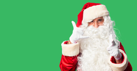 Middle age handsome man wearing Santa Claus costume and beard standing smiling doing talking on the telephone gesture and pointing to you. Call me.