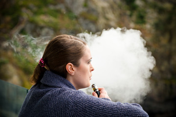 Young brunette caucasian woman smoking a vape ecigarette while standing on a balcony overlooking...