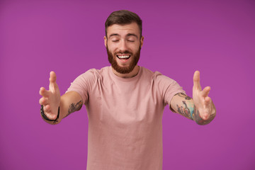 Pleasant looking happy brunette bearded male with tattooes standing over purple background with opened arms, going to give hug and keeping eyes closed