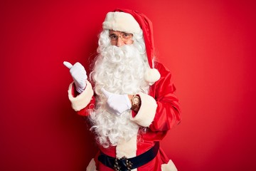 Middle age handsome man wearing Santa costume standing over isolated red background Pointing to the back behind with hand and thumbs up, smiling confident