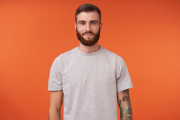 Attractive young bearded tattooed brunette man with trendy haircut standing over orange background,...