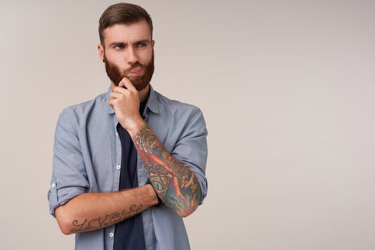 Severe young brunette unshaved male with tattooes holding his chin with raised hand and looking aside, frowning face and twisting his mouth while standing over white background