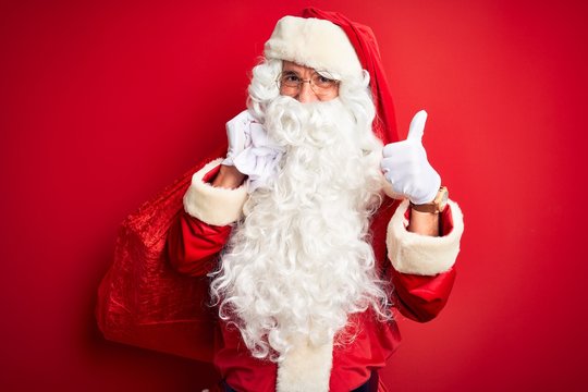 Middle age man wearing Santa costume holding sack with gifts over isolated red background happy with big smile doing ok sign, thumb up with fingers, excellent sign