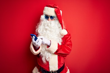 Senior man wearing Santa Claus costume using smartphone over isolated red background with a happy face standing and smiling with a confident smile showing teeth