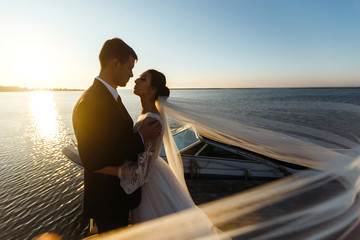 Pretty bride and stylish groom together on the bridge against the background of the boat. Newlyweds enjoy each other tenderly in the shadow of a flying veil. Together. Wedding. Love.