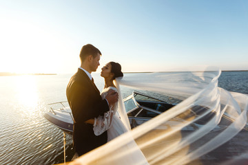 Pretty bride and stylish groom together on the bridge against the background of the boat. Newlyweds enjoy each other tenderly in the shadow of a flying veil. Together. Wedding. Love.