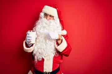 Senior man wearing Santa Claus costume holding glass of milk over isolated red background very happy pointing with hand and finger