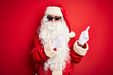 Obraz na płótnie Canvas Middle age man wearing Santa Claus costume and sunglasses over isolated red background smiling and looking at the camera pointing with two hands and fingers to the side.