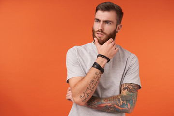 Pensive young tattooed brunette man in beige t-shirt and trendy accessories keeping hand on his lush beard and looking aside thoughtfully, isolated over orange background