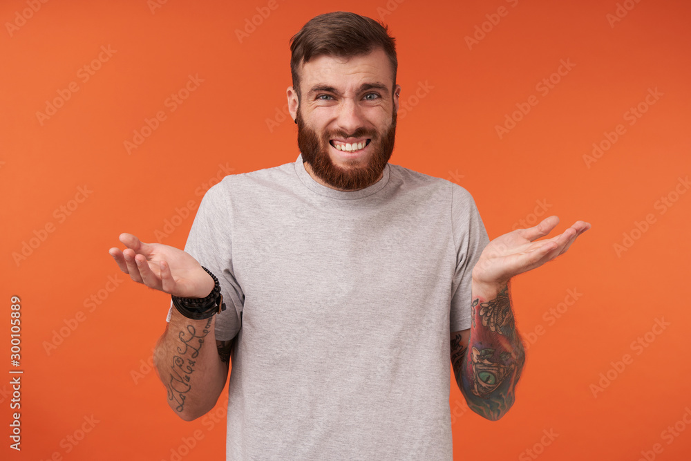 Wall mural Dissatisfied bearded brunette guy with trendy haircut looking at camera with pout and raising palms confusedly, wearing beige t-shirt and trendy accessories over orange background